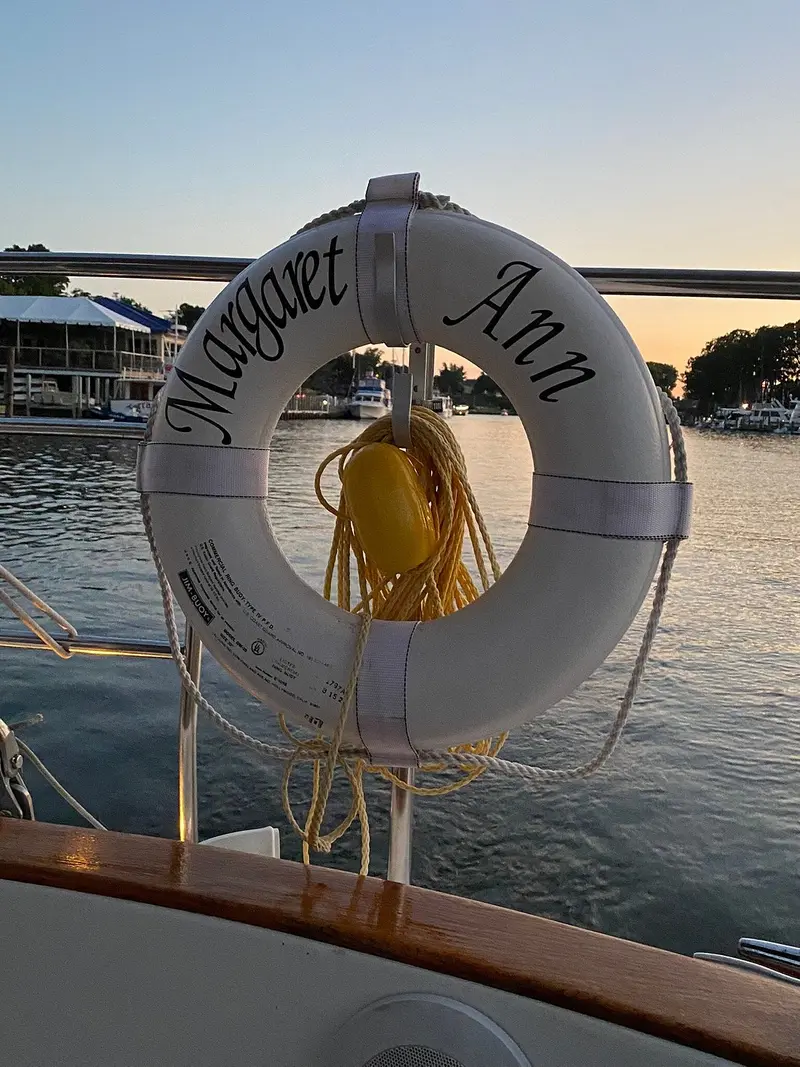A life ring marked "Margaret Ann" hanging on the boat’s rail at dusk, with docks and moored boats across the water behind it